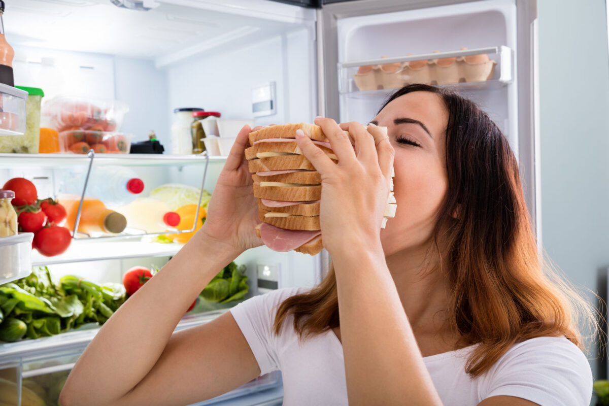 žena jede ogroman sendvič ispred otvorenog frižidera